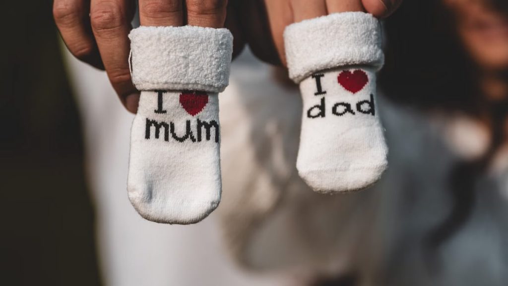 Close-up of parents holding baby socks indicating pregnancy announcement, tagged with 'I love mum' and 'I love dad'.